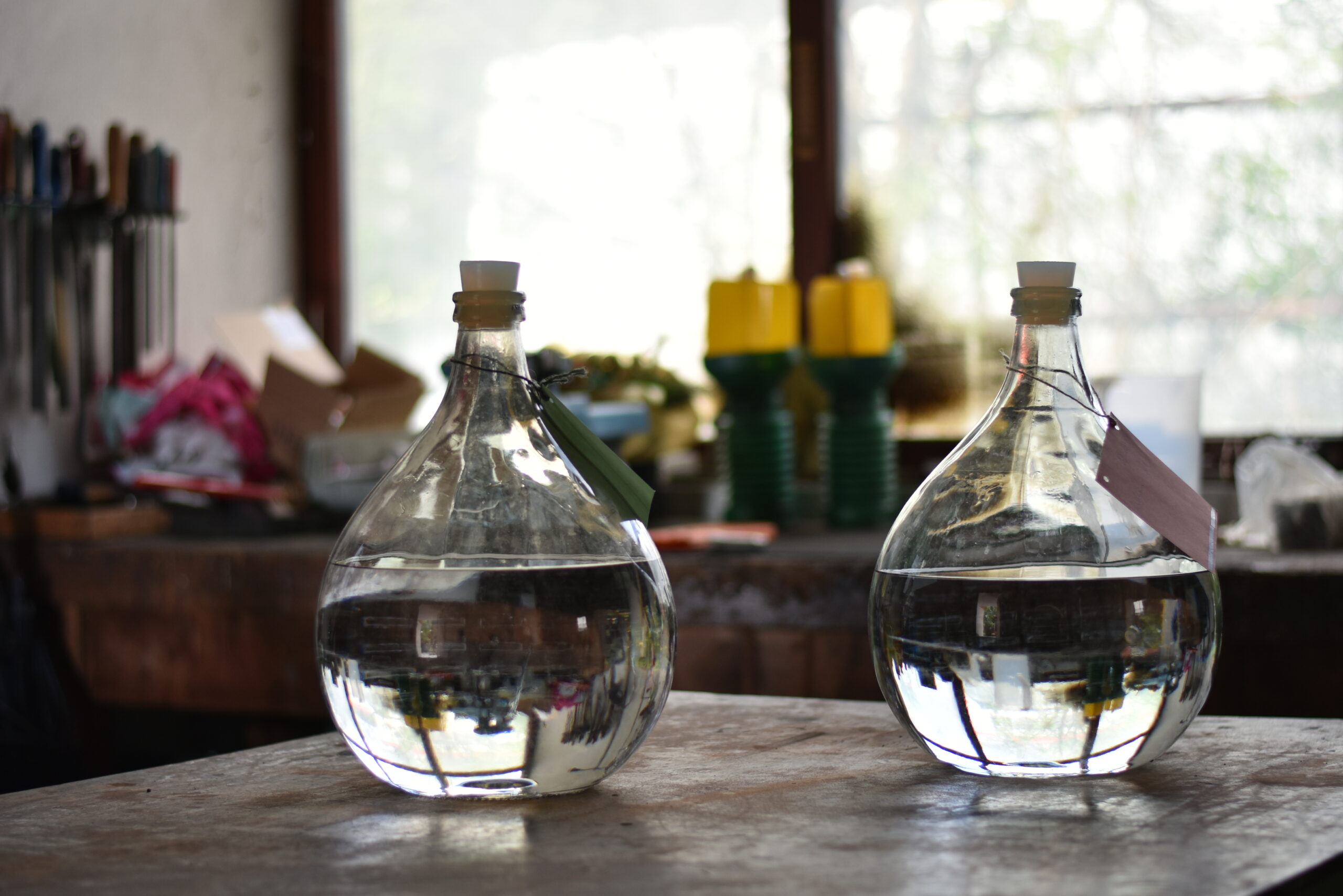 Edelbrand Auf einem Eisentisch stehen zwei große Glasballons. Diese sind mit klarer Flüssigkeit gefüllt. Im Hintergrund ist ein großes Fenster