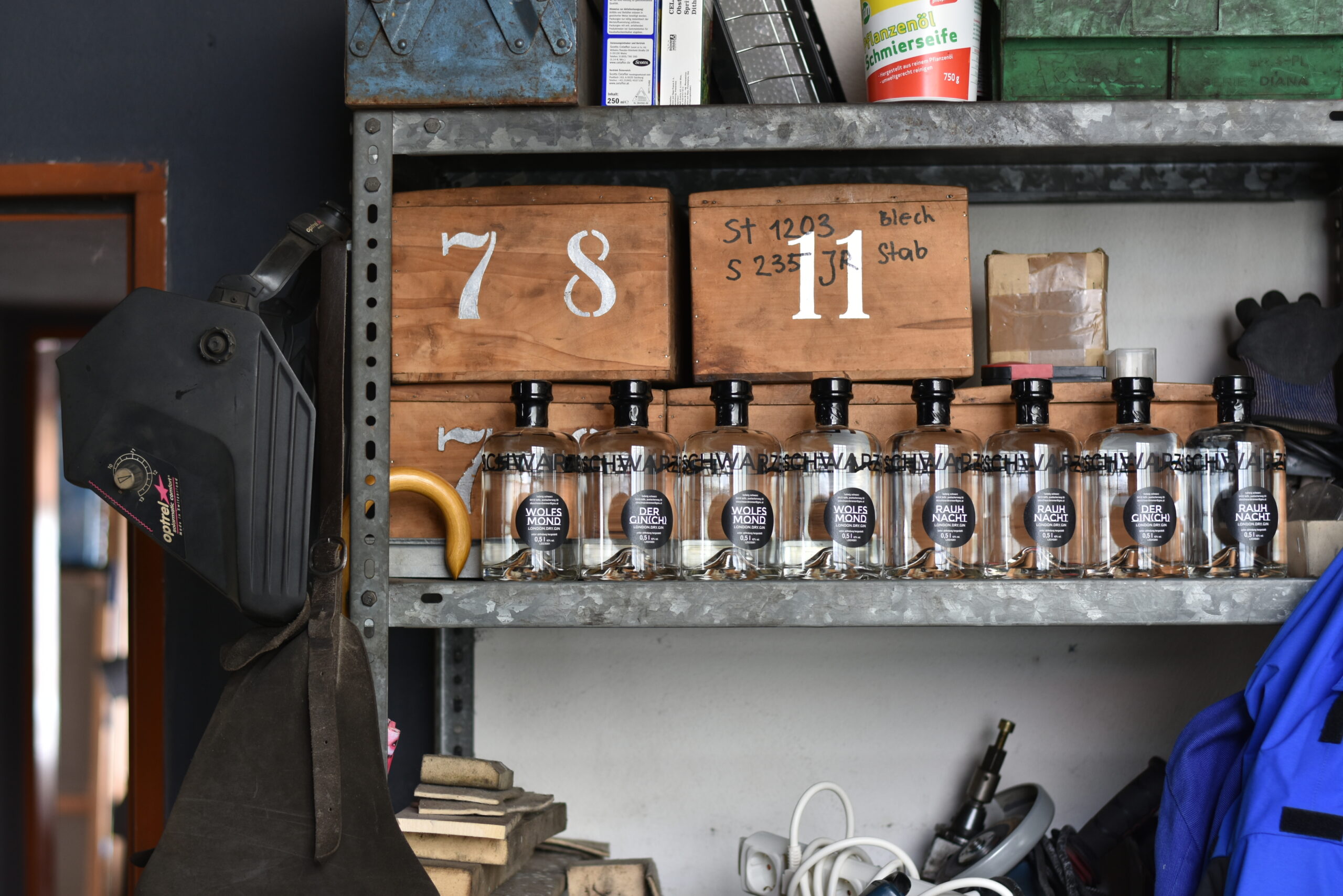 Gin Auf einem Schwerlastregal stehen nebeneinander Getränkeflaschen. Hinter den Flaschen sind Kisten und Werkzeuge in das Regal geschlichtet